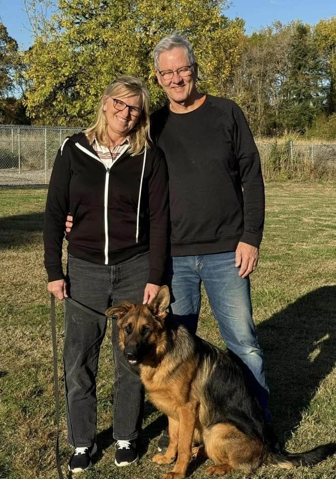 Robin and her husband standing in a field with their German Shepherd